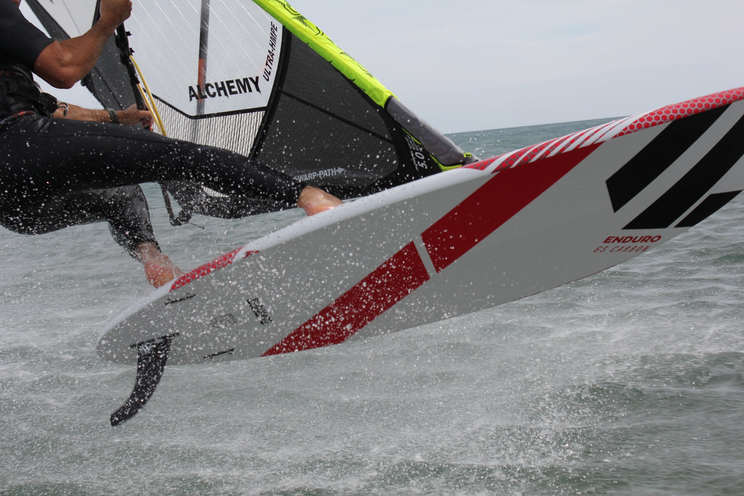 Person windsurfing on a wave with a sail branded 'Alchemy' and a red and white sail.